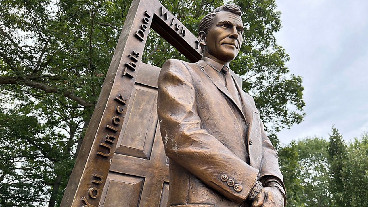 Low angle photo of statue of Rod Serling standing in a suit holding his hands in front of him. behind him is a crooked doorway with the door open ajar, with writing on the door frame