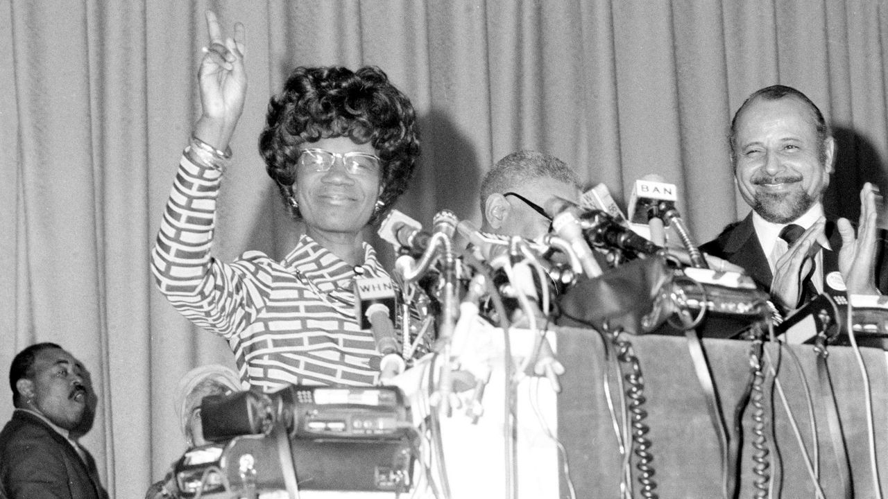 Rep. Shirley Chisholm is congratulated by Rep. Ronald V. Dellums after her formal announcement in New York, Jan. 25, 1972, that she will seek the Democratic presidential nomination. She is standing behind a podium with many mics attached and holding her arm up with a "peace " sign.