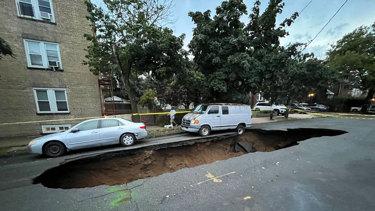 A large sinkhole on a street in the Bronx is widening and a white van and a silver sedan are about to fall into the hole. 