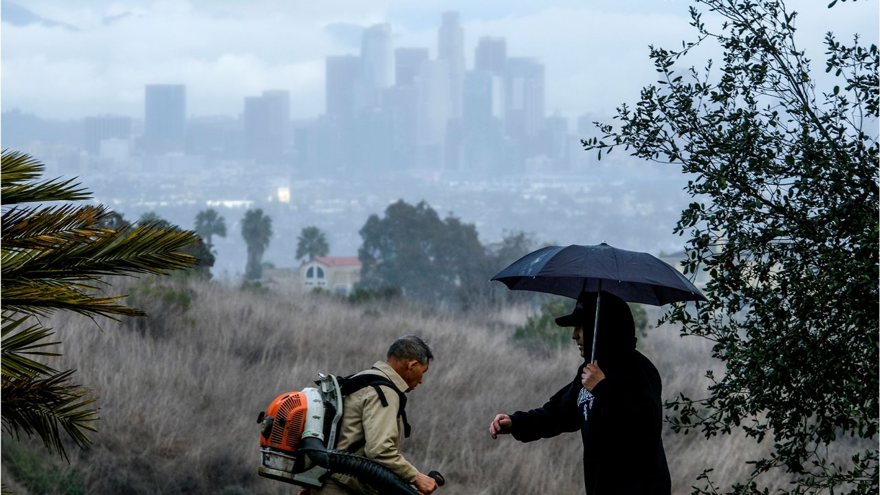 A man with an umbrella walks by a worker in Kenneth Hahn State Recreation Area as the Los Angeles skyline in background on Friday, Dec. 24, 2021. (AP Photo/Ringo H.W. Chiu)