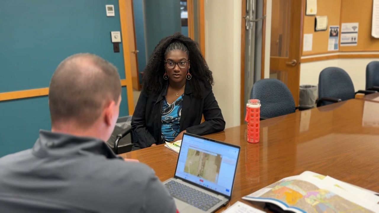 intern in conference room, wearing black blazer, talks with man sitting at end of table, who has a laptop open