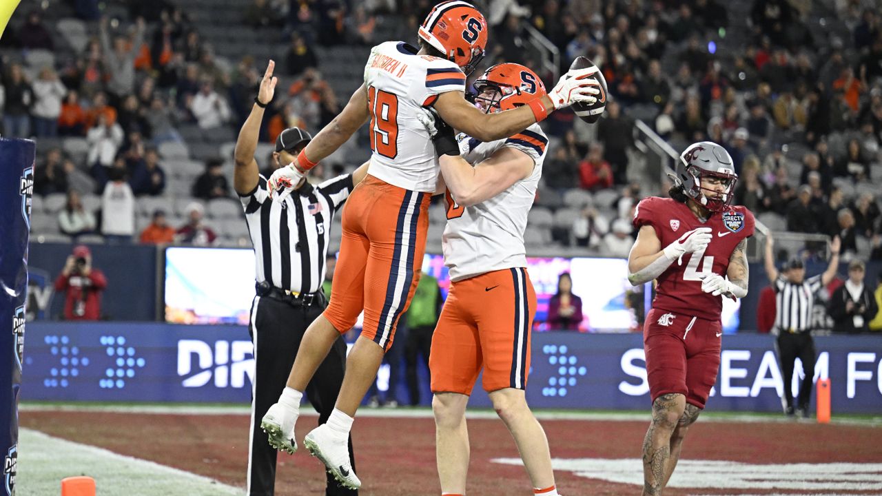 syracuse football player lifts teammate, holding ball in end zone, to celebrate touchdown as washington state player jogs away in background