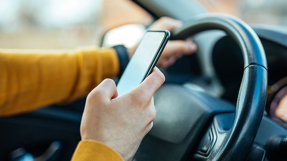 A person holds their phone in one hand while their other hand is on the wheel of their car. (Getty Images)