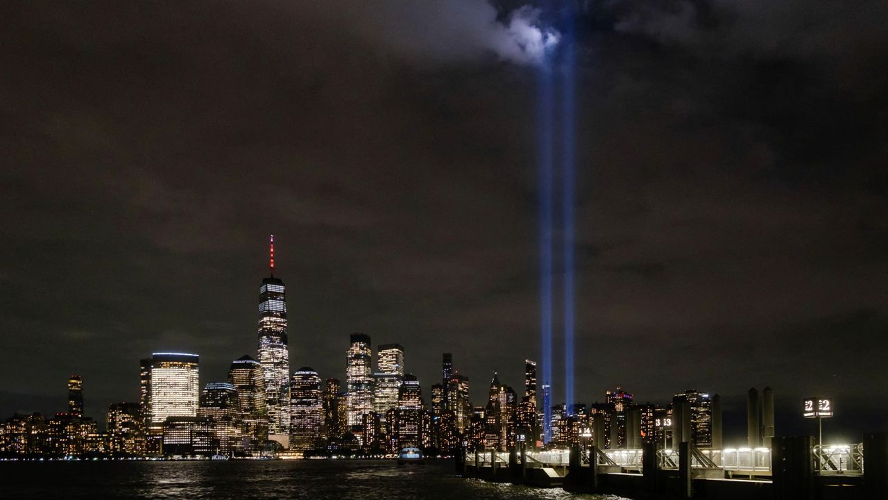 Twin beams of light commemorating the fallen twin towers of the World Trade Center are seen against a night sky with water in the foreground and buildings surrounding the light beams