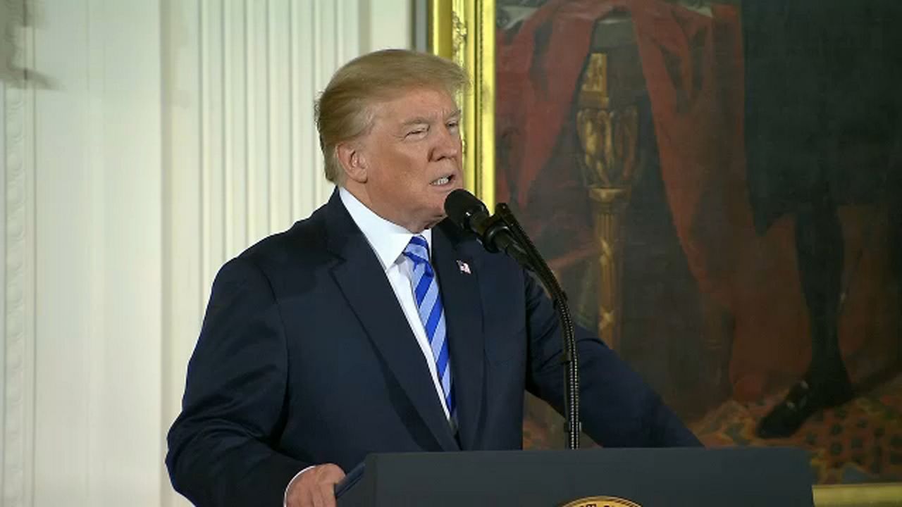 President Donald Trump, wearing a black suit, a white dress shirt, and a blue-striped tie, speaks into a black microphone that juts out of the center of a black lectern, which he grips with his right hand. A painting is behind him, along with white wallpaper.