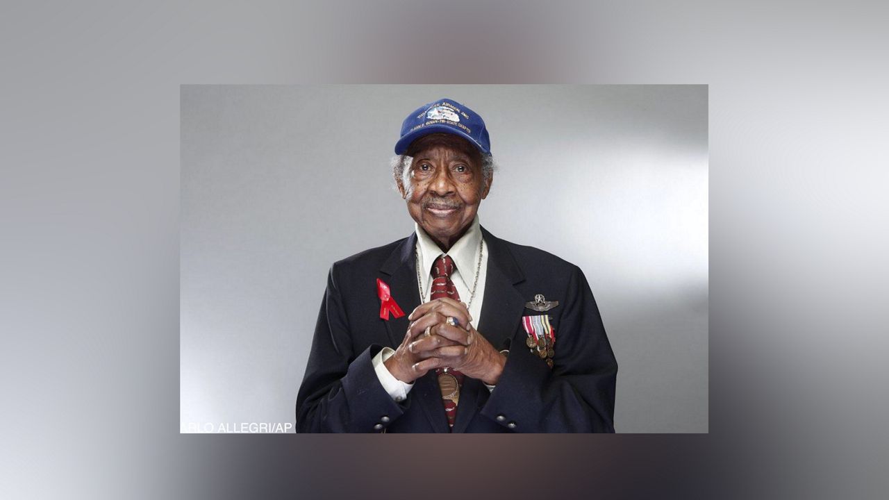 Floyd Carter Sr., wearing a black suit, a white dress shirt, a red tie, and a blue Tuskegee Airman hat. Medals and a red bow are pinned to his suit jacket.