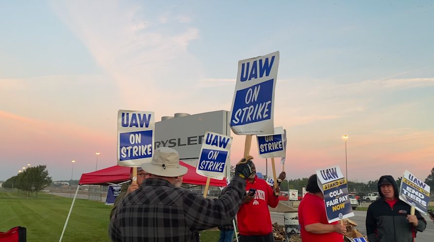 Workers stand outside an automobile assembly plant with strike signs as the sun rises behind them.