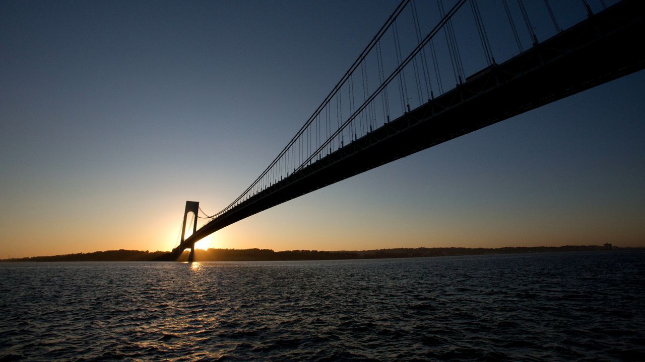 A photograph of the Verrazzano-Narrows Bridge from the Brooklyn side as the sun sets over the other end of the bridge.