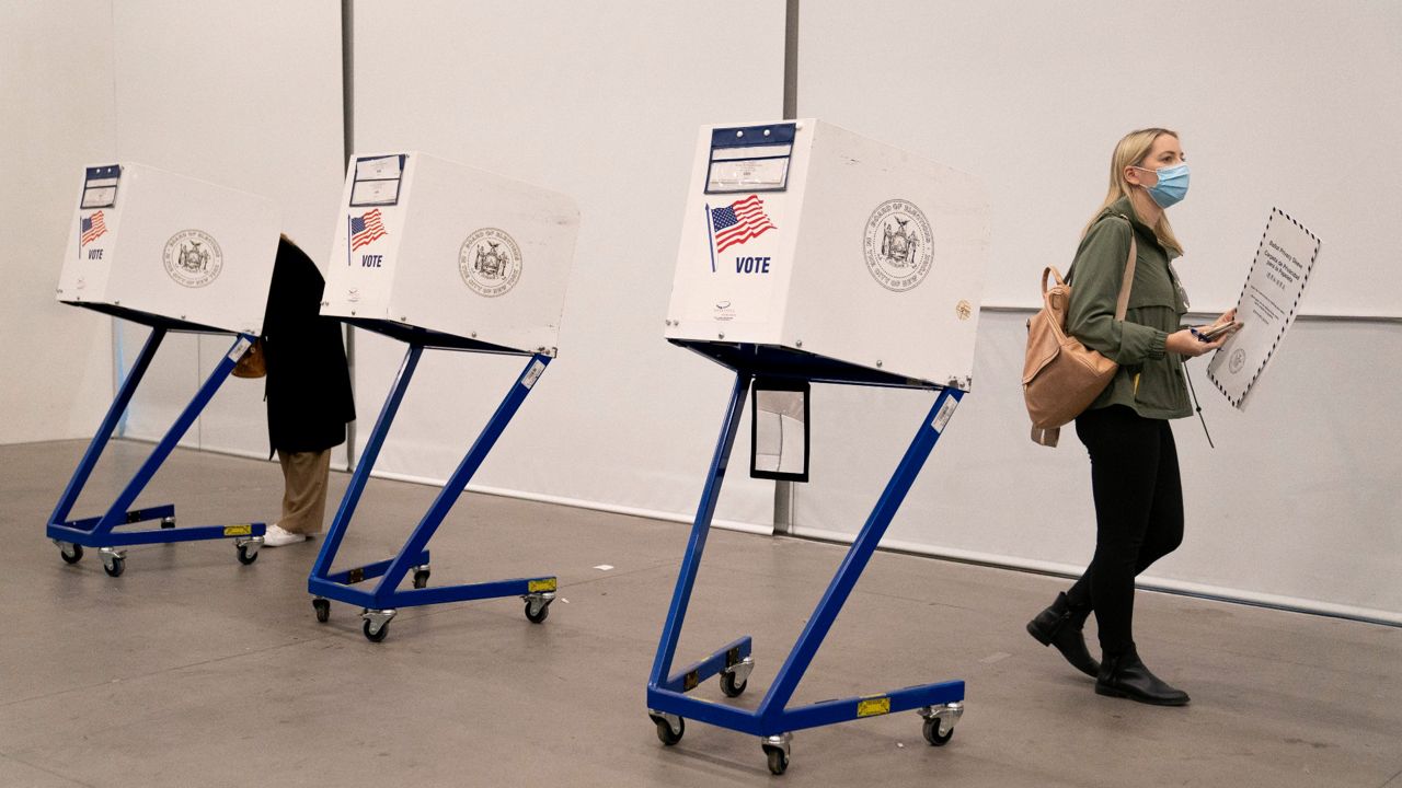 A woman carries her ballot folder while walking away from three voting machines set up in a room with concrete floors and a white wall. 