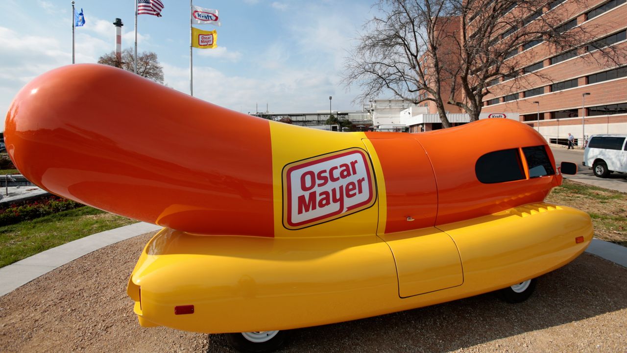 Oscar Mayer wienermobile parked outside of a building. Flags, including an American flag and an Oscar Mayer flag are seen in the background. 