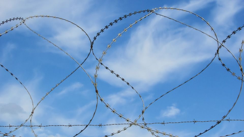Zoomed in photo of barbed wire against a blue sky with clouds. 