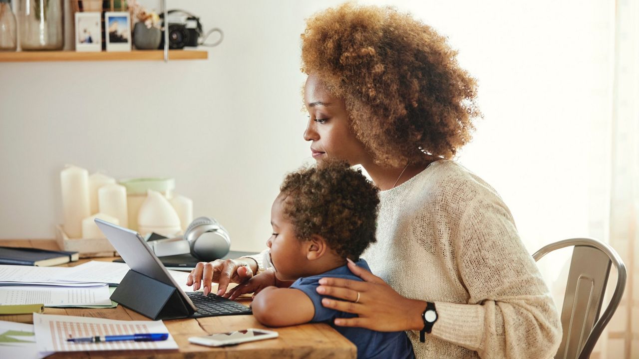A black woman with an infant on her lap sitting at table strewn with office supplies and both are looking at a tablet computer.