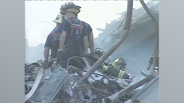 People wearing blue FDNY shirts and black FDNY hats, while wearing white masks, stand near a pile of rubble.