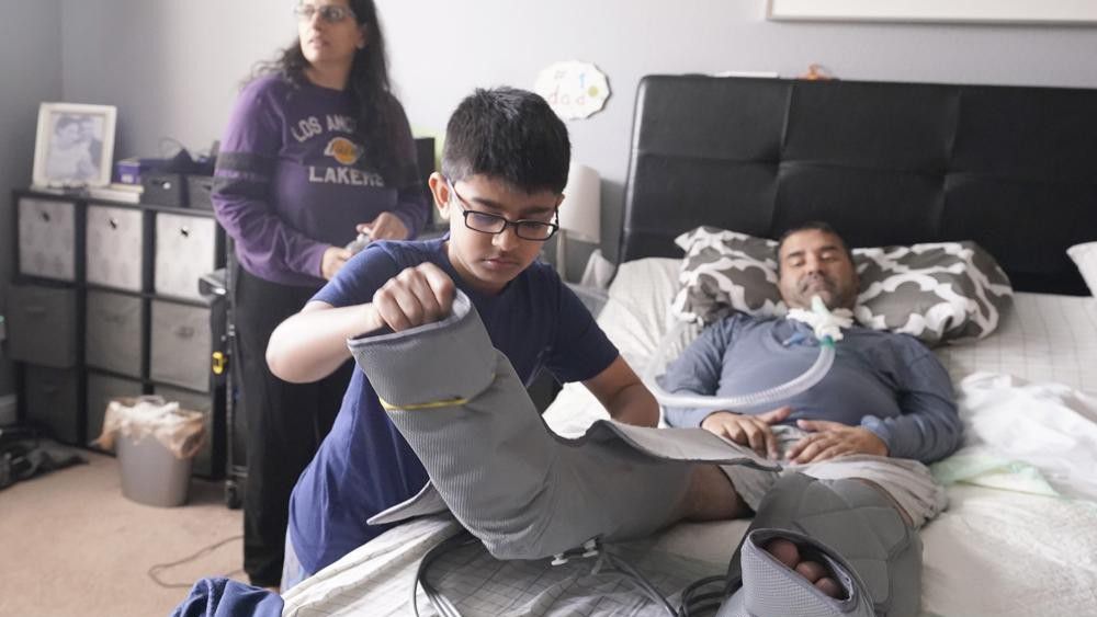 Ronan Kotiya, 11, removes a compression leg sleeve from his father Rupesh Kotiya as his mother Siobhan Pandya looks at their home in Plano, Texas, Sunday, April 10, 2022. (AP Photo/LM Otero)