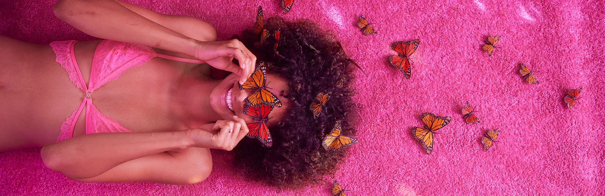 Model in pink lace bralette laying on pink grass surroded by butterflies