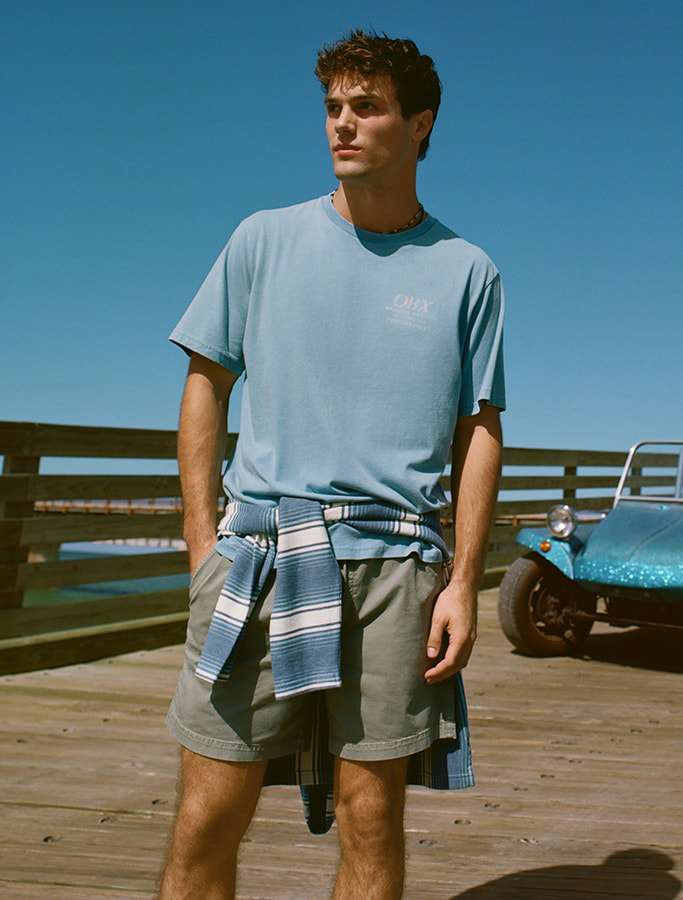 model in a blue t-shirt with grey shorts and a striped hoodie wrapped around his waist standing on a boardwalk