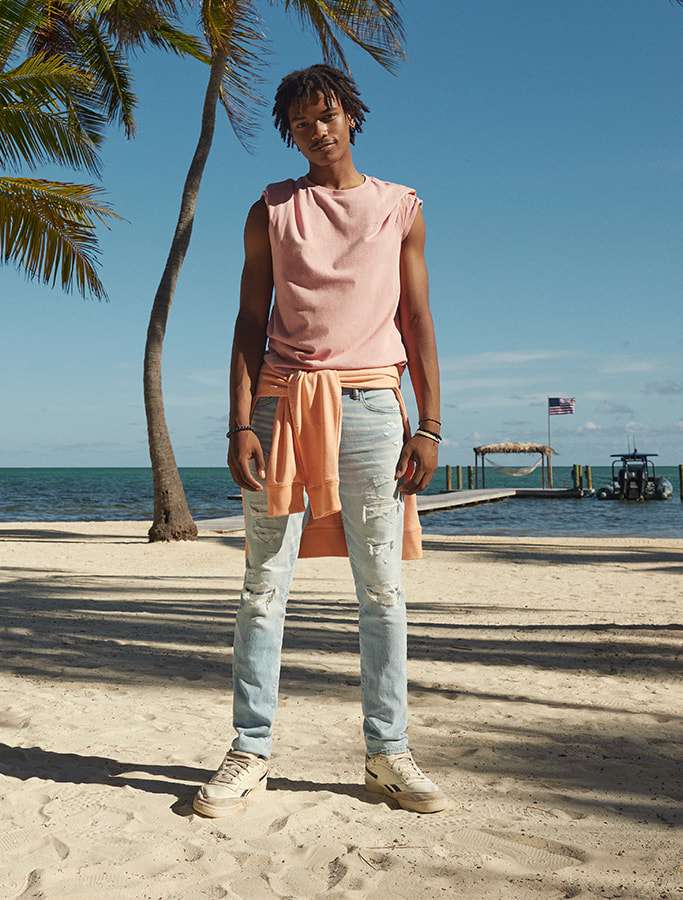 model on the beach in light blue jeans, a pink tee with a light orange hoodie wrapped around his waist and in camel colored boots