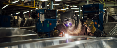 A welder wearing a protective helmet and gloves is working on a Lowe Boat at the Lowe Factory. The welder uses a welding torch and filler rod, creating bright sparks as the metal is fused together. The background shows various machinery, equipment, and other metal structures under fluorescent lighting in the workshop.