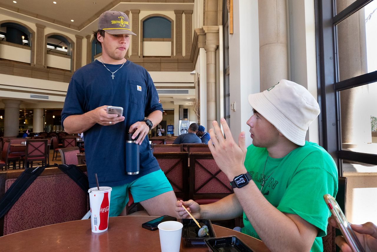 Troy standing and talking to Logan sitting at a table