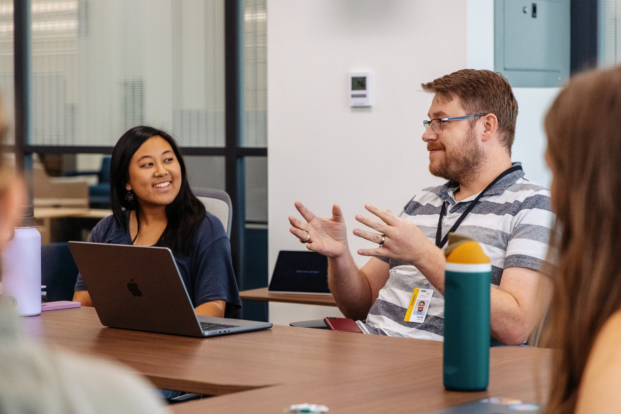 Person sitting at table in an open floor plan working on laptop with others.