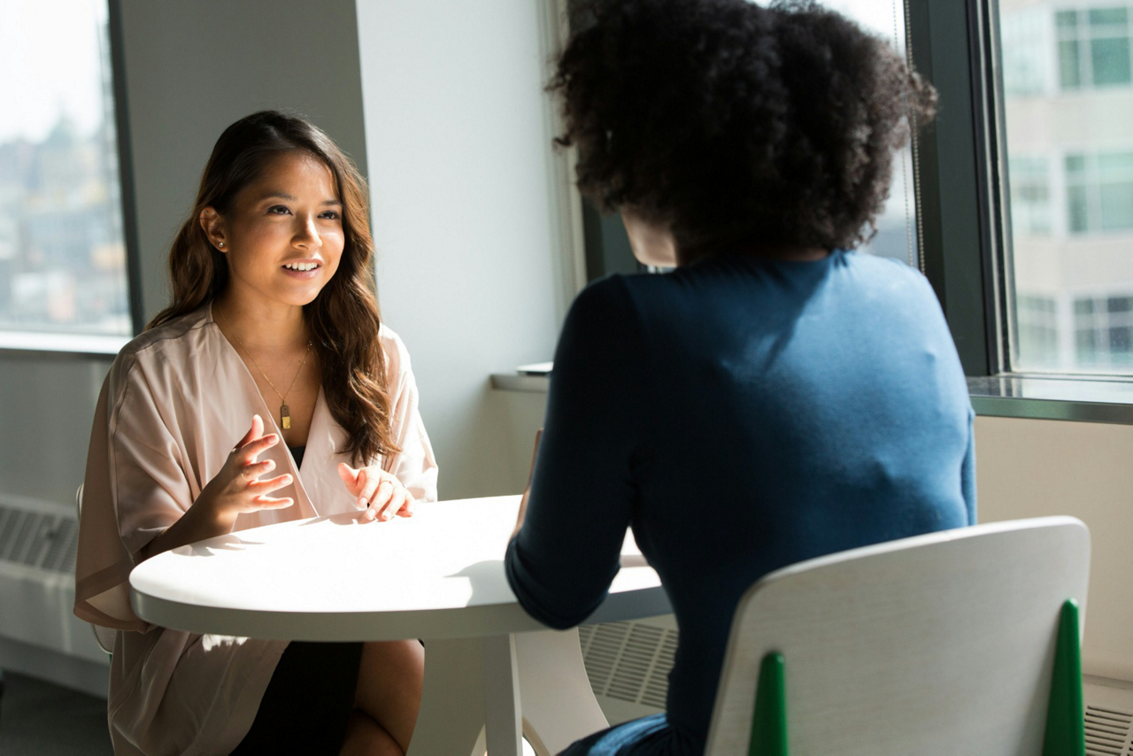 Two women talking at a table beside a window