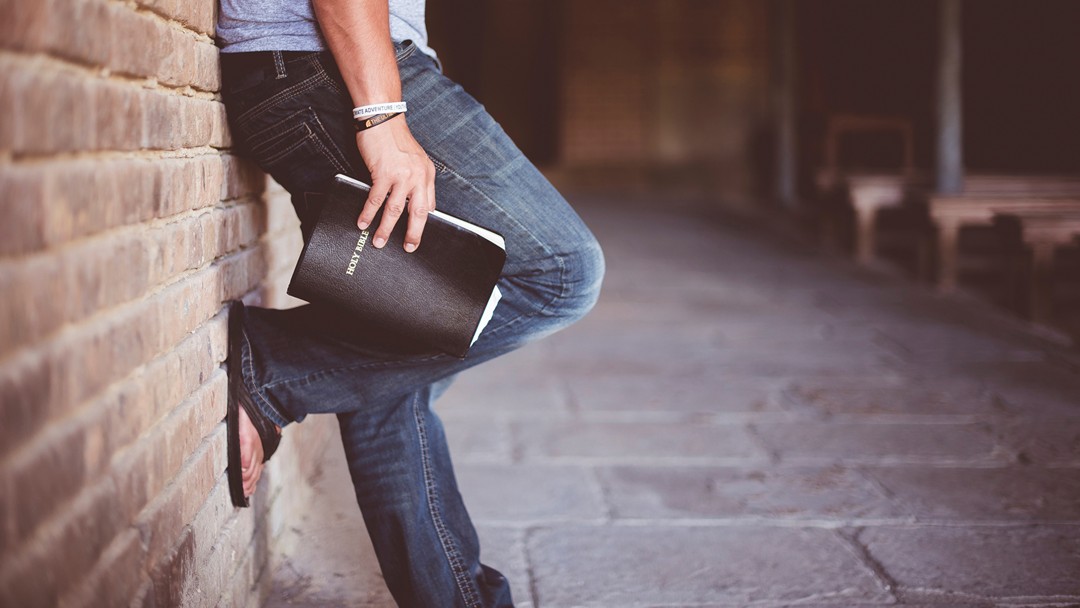 Man holding bible leaning against a brick wall.