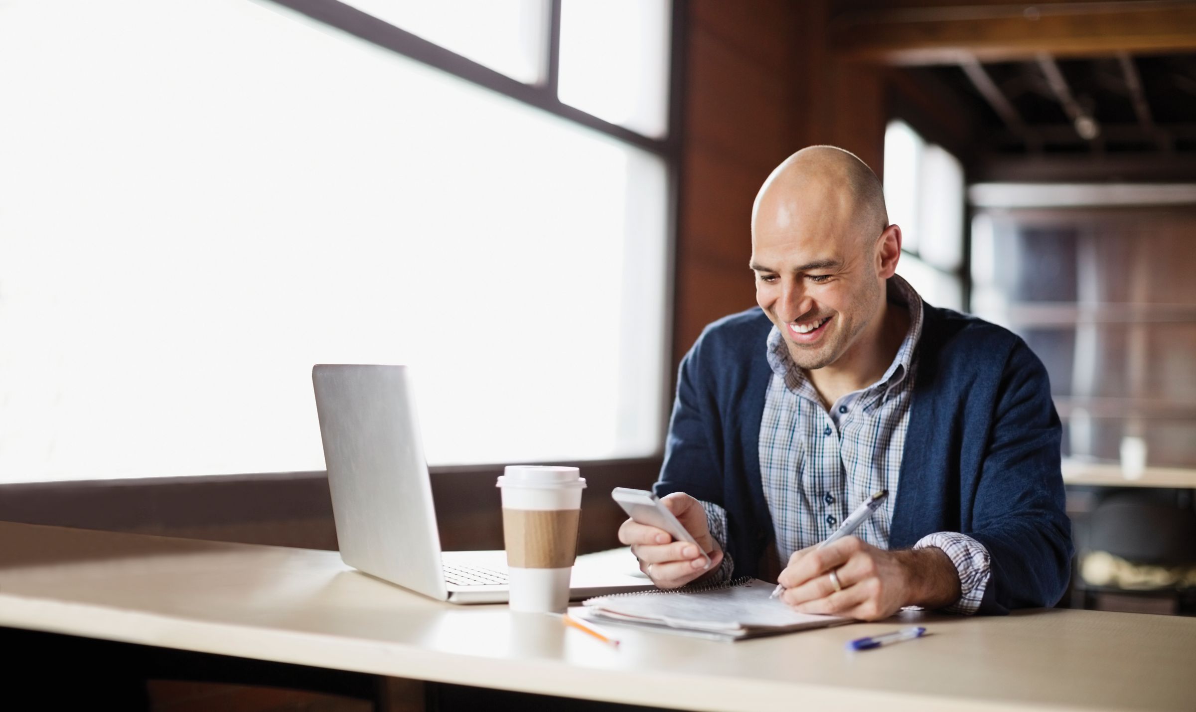 a man is sitting at a desk smiling while looking at his phone which is placed alongside a laptop