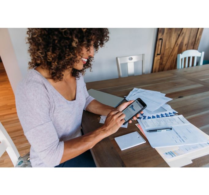 A woman sits at her dining room table with laptop and financial reports doing her monthly budget. She is smiling at the ease of use as she works on her smart phone banking app to do monthly finances, pay taxes and save money for the future.