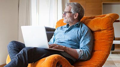 Gray haired man sitting in an orange chair holding a laptop 