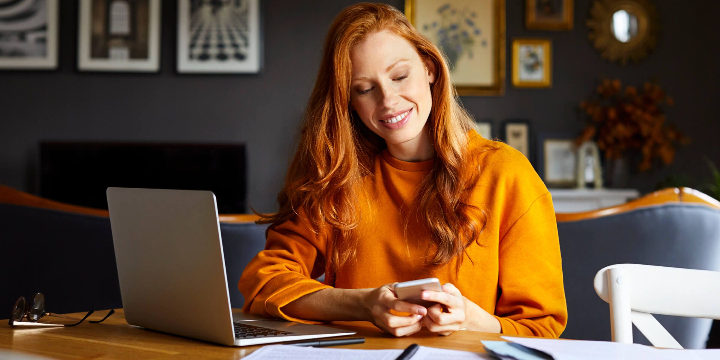 red headed lady sitting at table with laptop looking at phone 