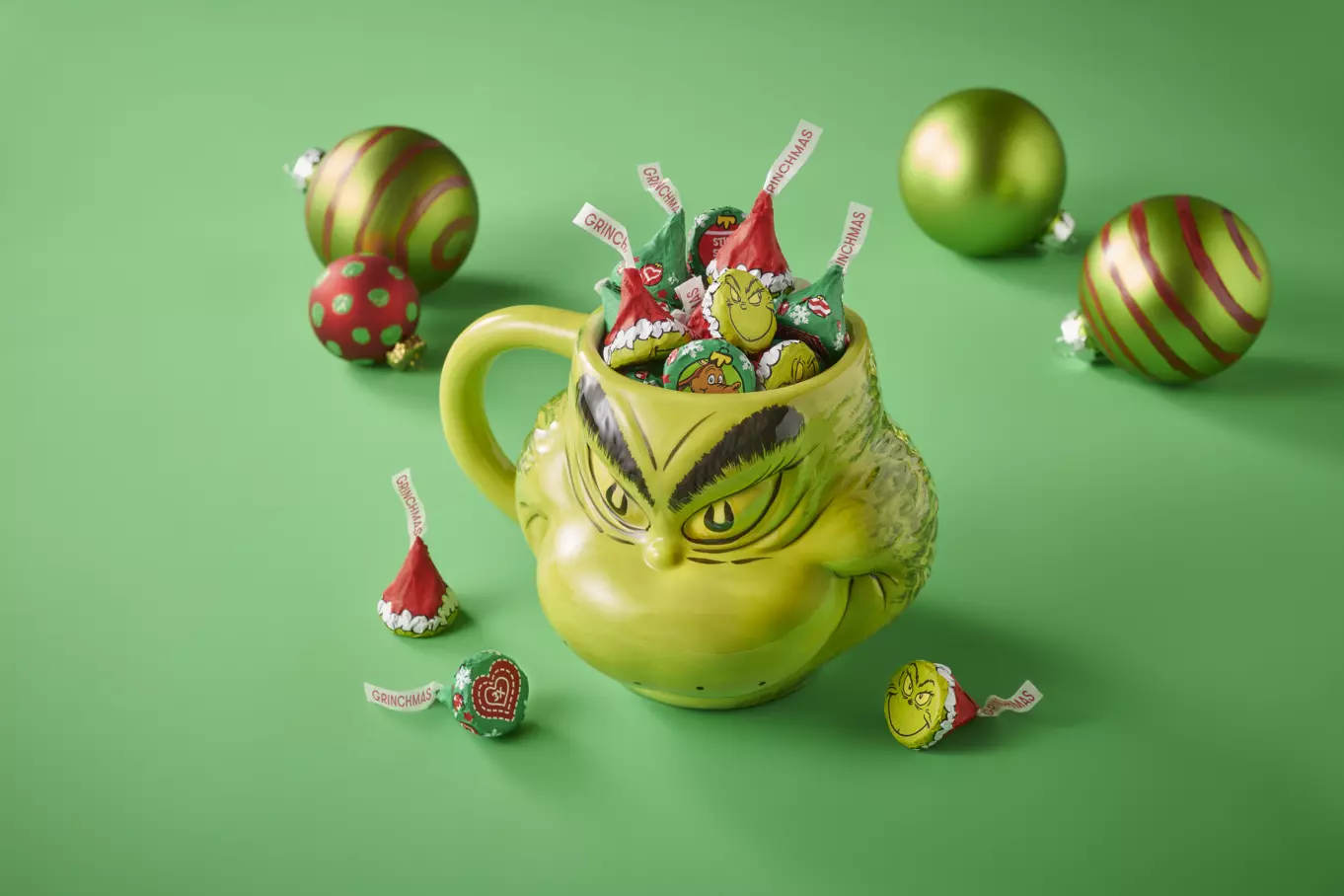 HERSHEY'S KISSES Grinch® Foils Candy inside Grinch themed mug surrounded by ornaments