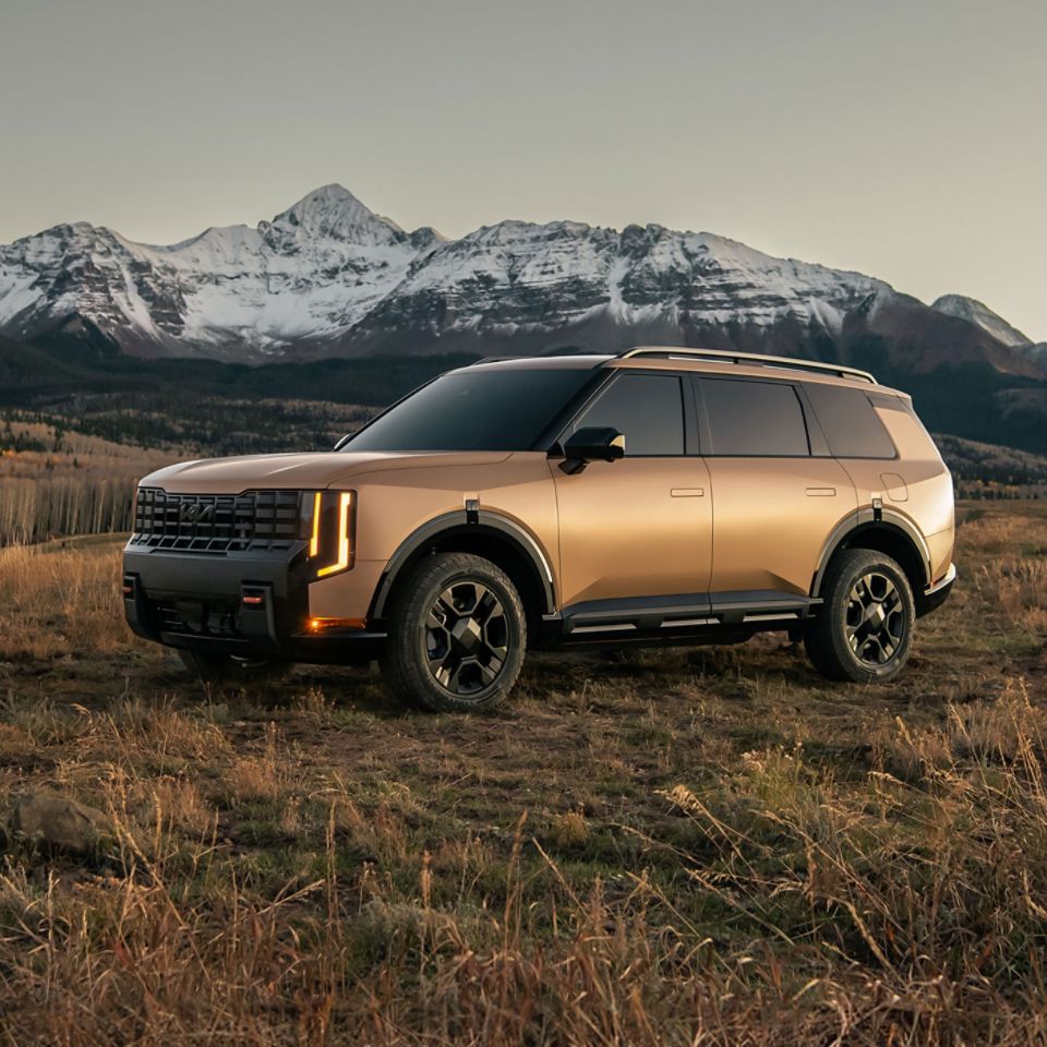 Driver-side front angle view of the 2027 Kia Telluride SUV in a shining golden tan color, with dark interior, black highlights, and glowing orange headlights. It looks like a natural part of the rugged nature scene around it; the vehicle is parked in a field, with imposing white-capped mountain peaks in the distance of the background.
