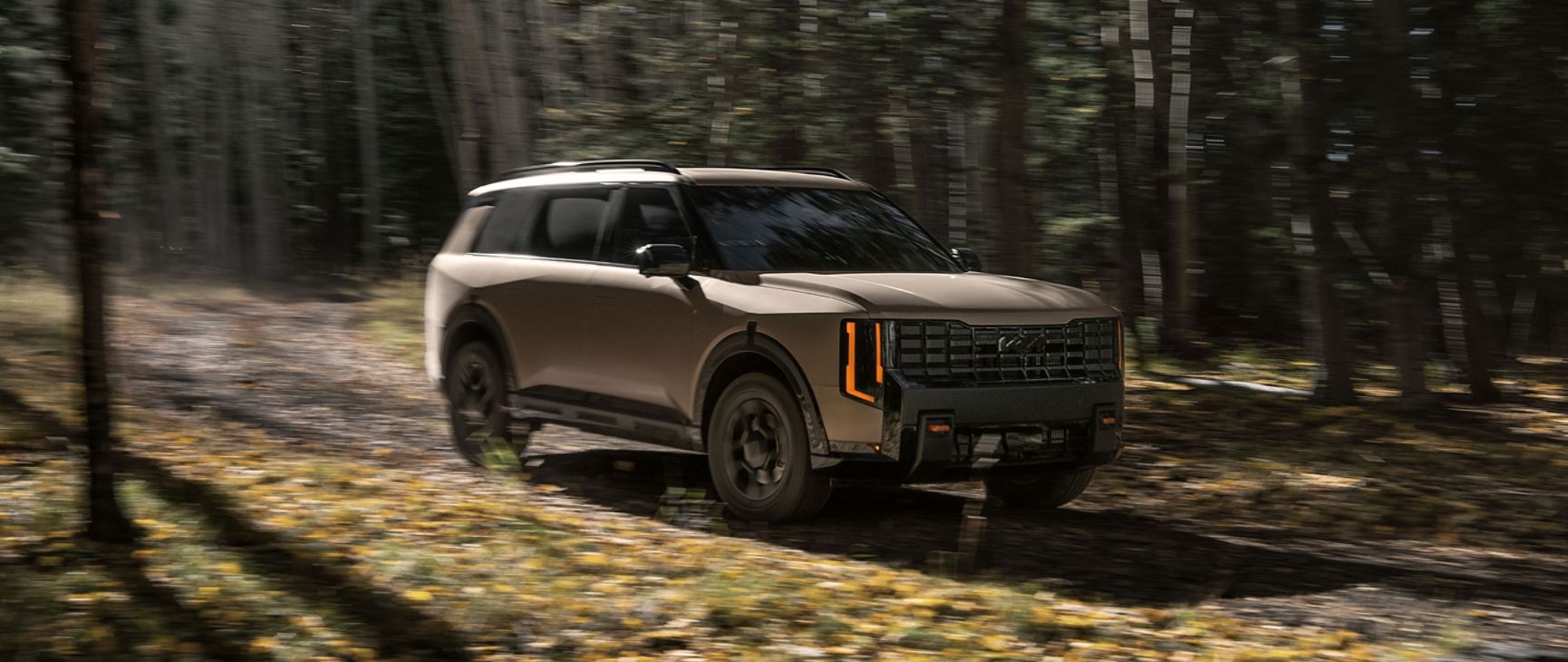 A dynamic shot of the Kia Telluride in tan as it speeds along a forest path that highlights the front and passenger side. Leaves and dirt scatter across the trail beneath it, while dense trees blur into the background.