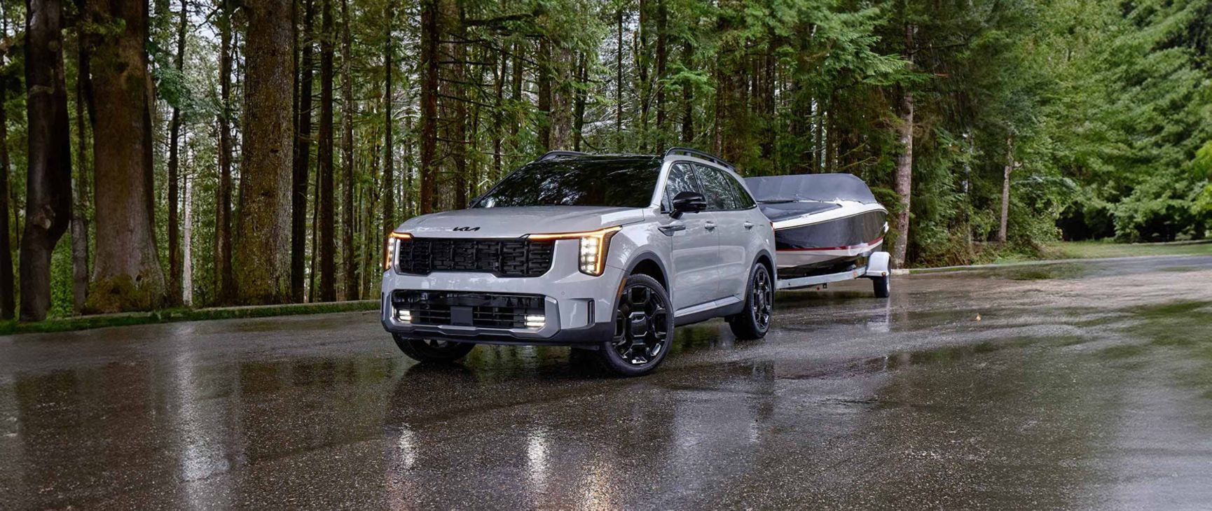 A matte gray Kia Telluride on a rainy road, framed by tall, lush trees. The SUV’s bold front grille is fully visible as it securely tows a boat hitched to the rear, emphasizing its strength and capability.