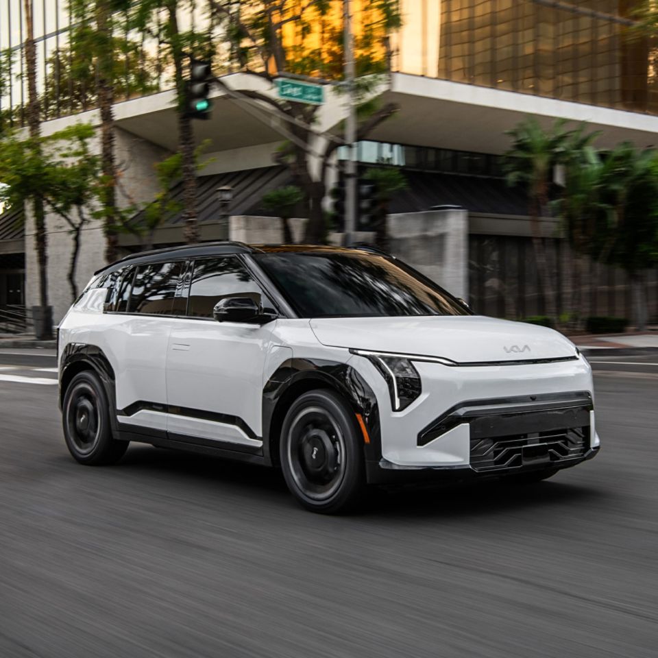 The 2027 Kia EV3 electric SUV GT-Line trim in white driving down an urban road with trees and interesting architecture.