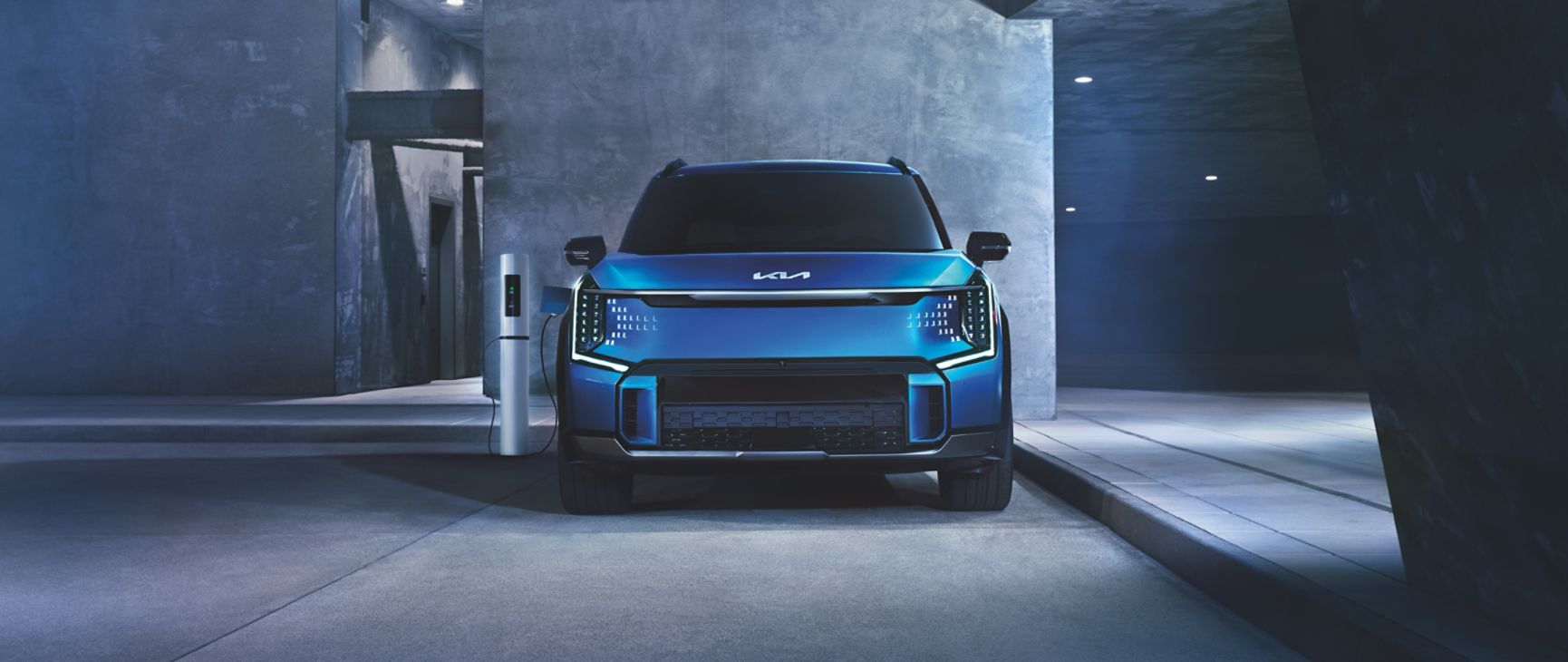 2024 Kia EV9 in blue, front view, plugged into a Level 2 charger in an at-home garage