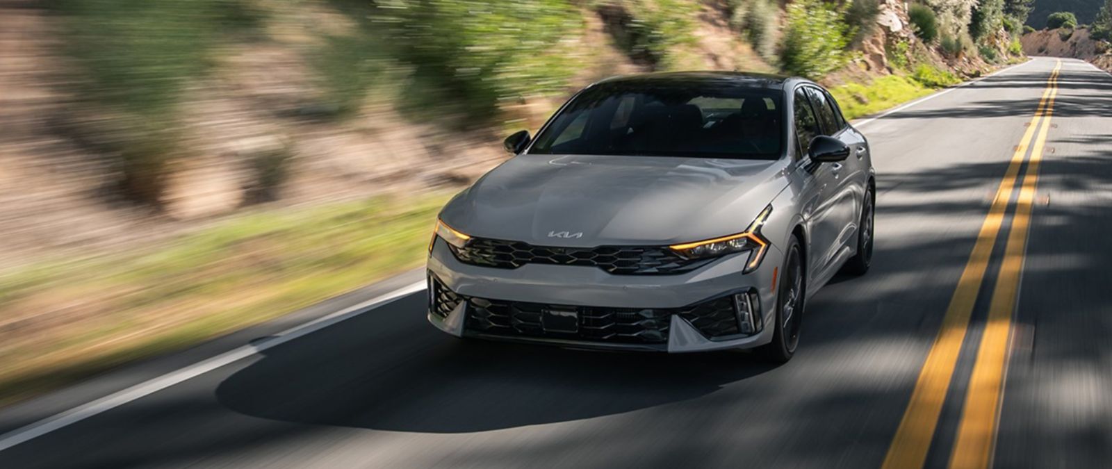 Front driver-side view of the 2025 Kia K5 driving by a blurred mountain background