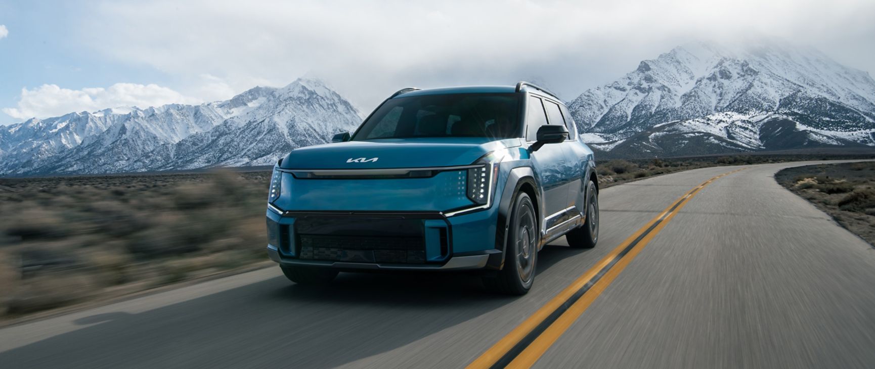 Blue 2026 Kia EV9 driving along the highway with snowy mountains in the background