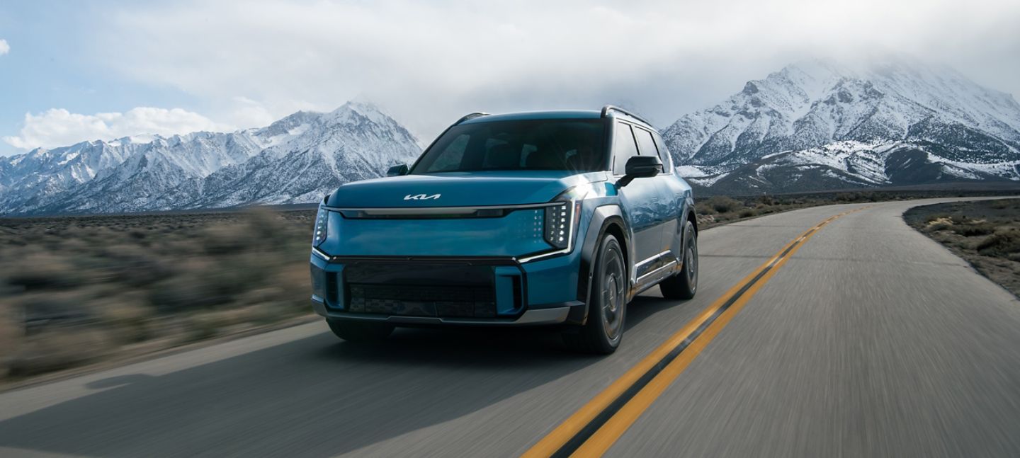 Blue Kia EV9 three-row electric SUV driving on a desert road with snowy mountains behind it