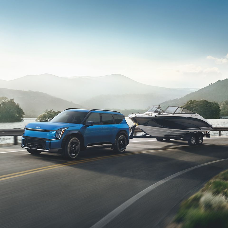 Side view of a blue 2026 Kia EV9 towing a boat on the highway