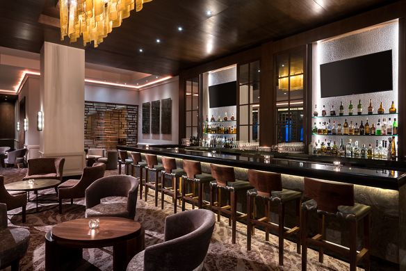 Lounge tables beneath a sculptural lighting fixture next to a bar with stools and wall-mounted flat-screen TVs