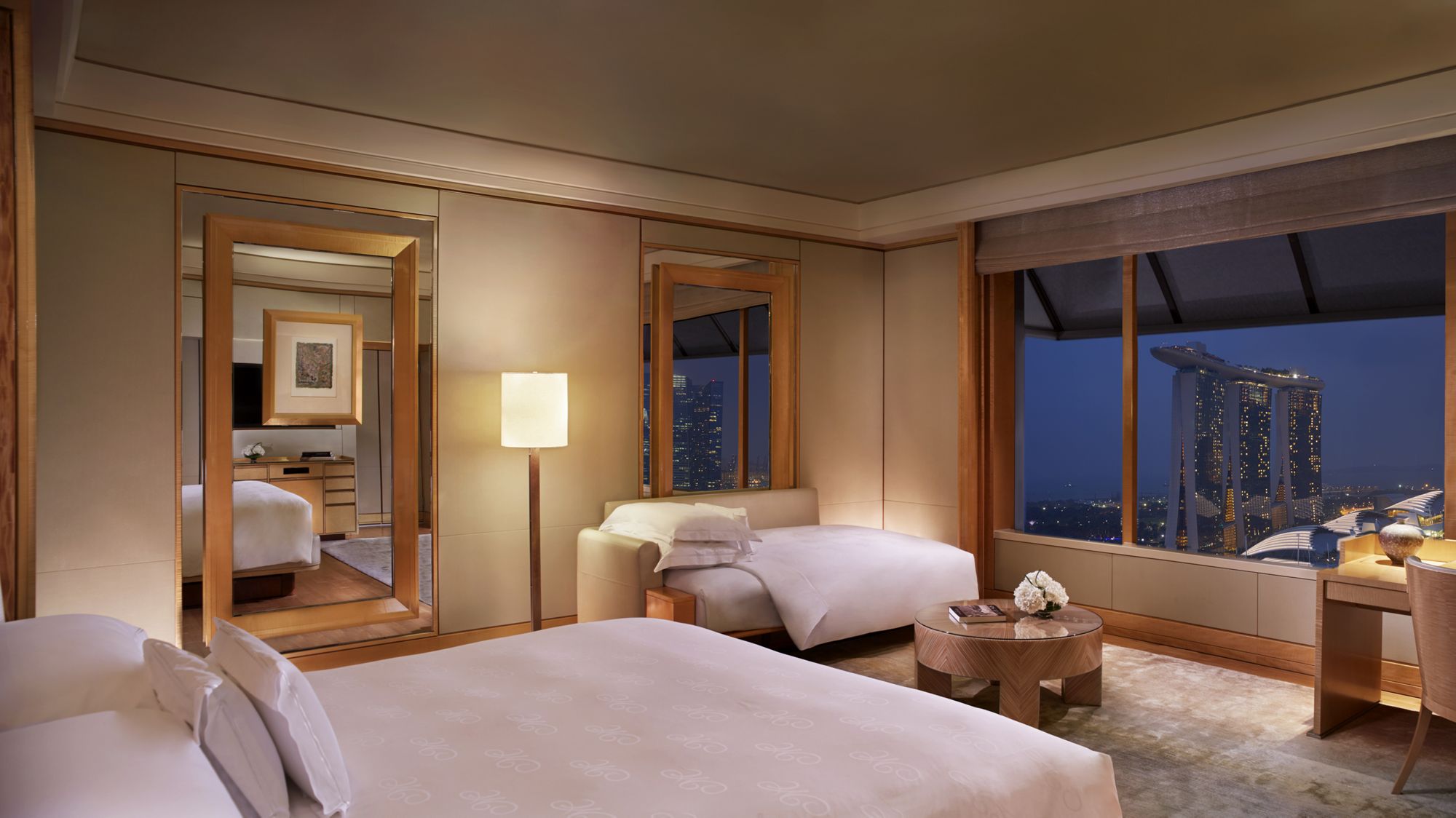 Guest room outfitted in honey-colored wood and carpet with wall-to-wall windows overlooking city lights in the evening