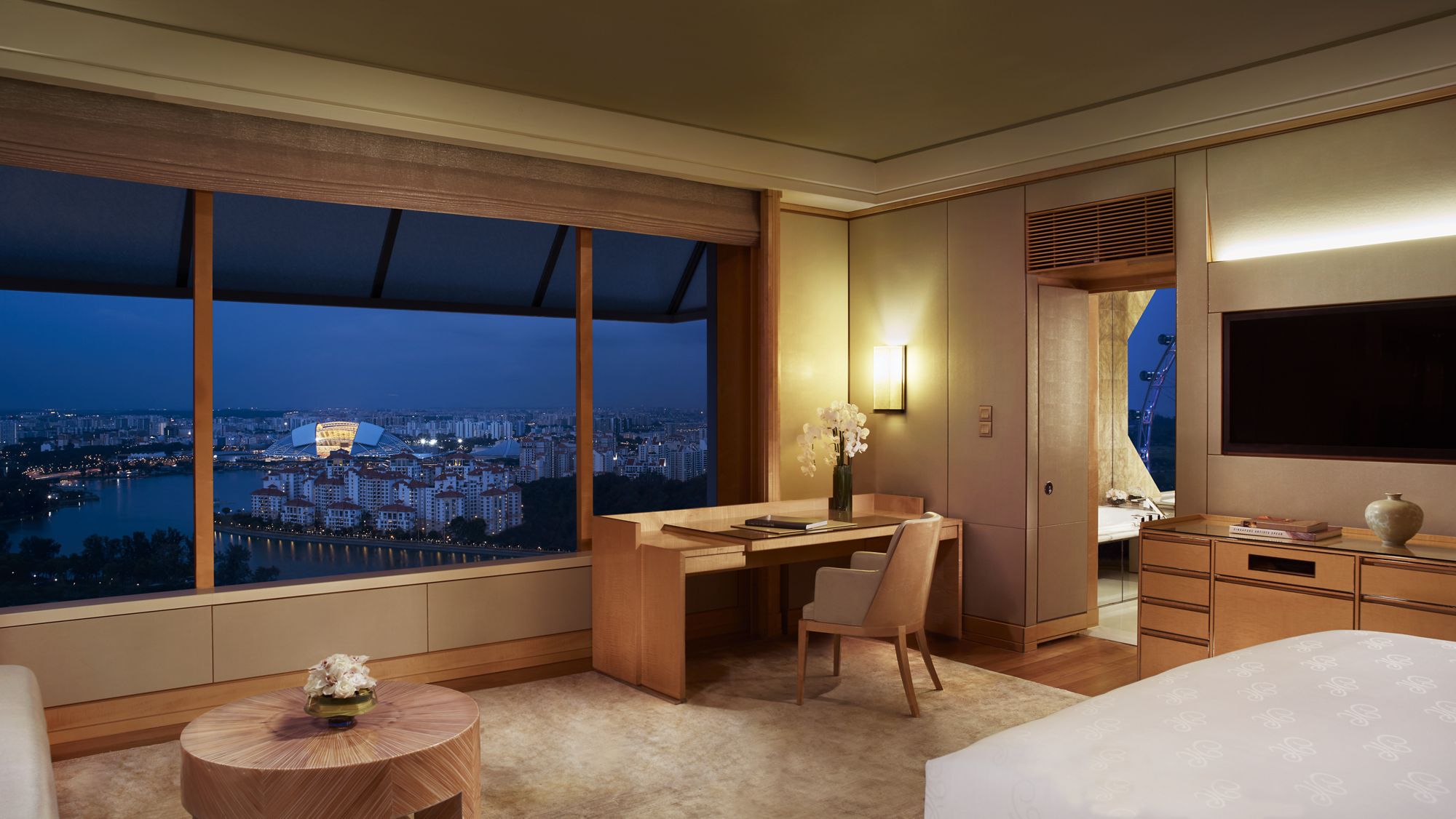 Guest room outfitted in honey-colored wood and carpet with wall-to-wall windows overlooking the city lights in the evening