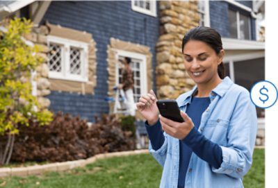 A person looking at a smart phone while a person stands on a ladder to paint exterior trim.