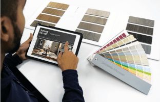 A person sitting at a table using an iPad looking at room paint colors with a color fan deck and color samples on the table.