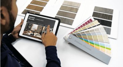 A person sitting at a table using an iPad looking at room paint colors with a color fan deck and color samples on the table.