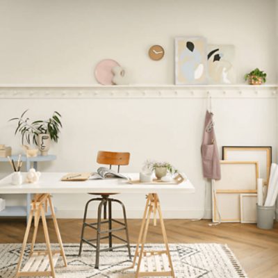 A crafting table with a light wood trestle base and a white top sits on a white tasseled rug with black geometric patterns. A stool with a short wood back is pulled up to the table. The wall behind is painted Alabaster SW 7008 an has a small clock, a few pieces of abstract artwork and a green leafy plant on a ledge midway up the wall. Blank canvases sit on the floor to the right of a cart with supplies.