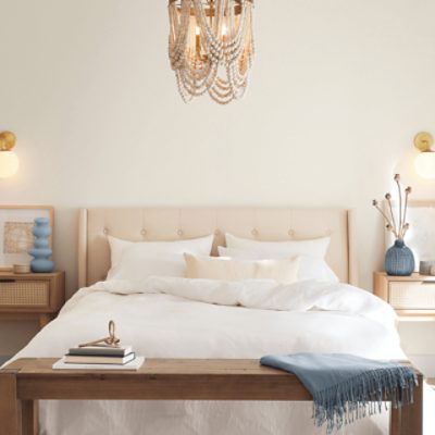 A bedroom with walls painted in a light tan color, chandelier bead light fixture, bed with white sheets and a wooden bench a the foot of the bed.