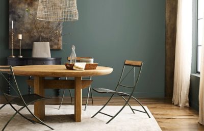 A dining area with wood and metal chairs around a light wood table with a cream rug and green-gray walls.