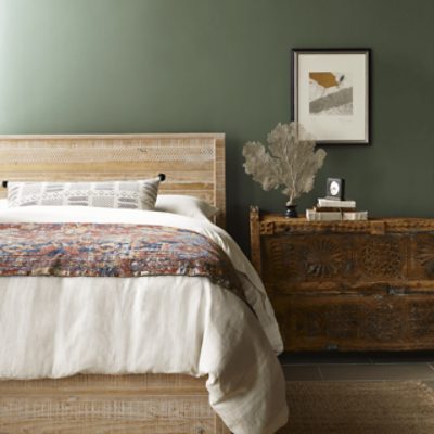 A bedroom with a bed with a wooden frame next to a wooden night stand against an olive green wall.